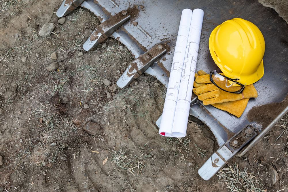 A yellow hard hat, rolled blueprints, work gloves, and safety glasses rest on the bucket of a construction excavator at a dirt-covered site during site preparation.